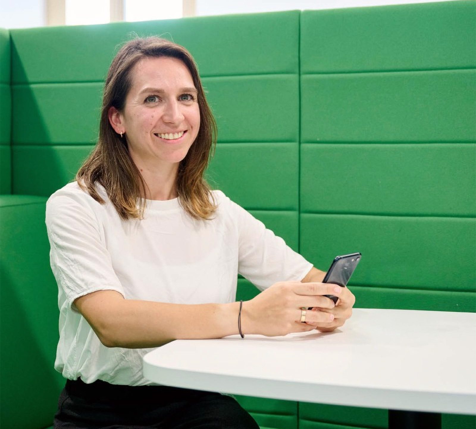 Eine Person sitzt an einem Tisch mit einem Smartphone in der Hand, in einem Raum mit grünen Wänden und einer modernen Atmosphäre.