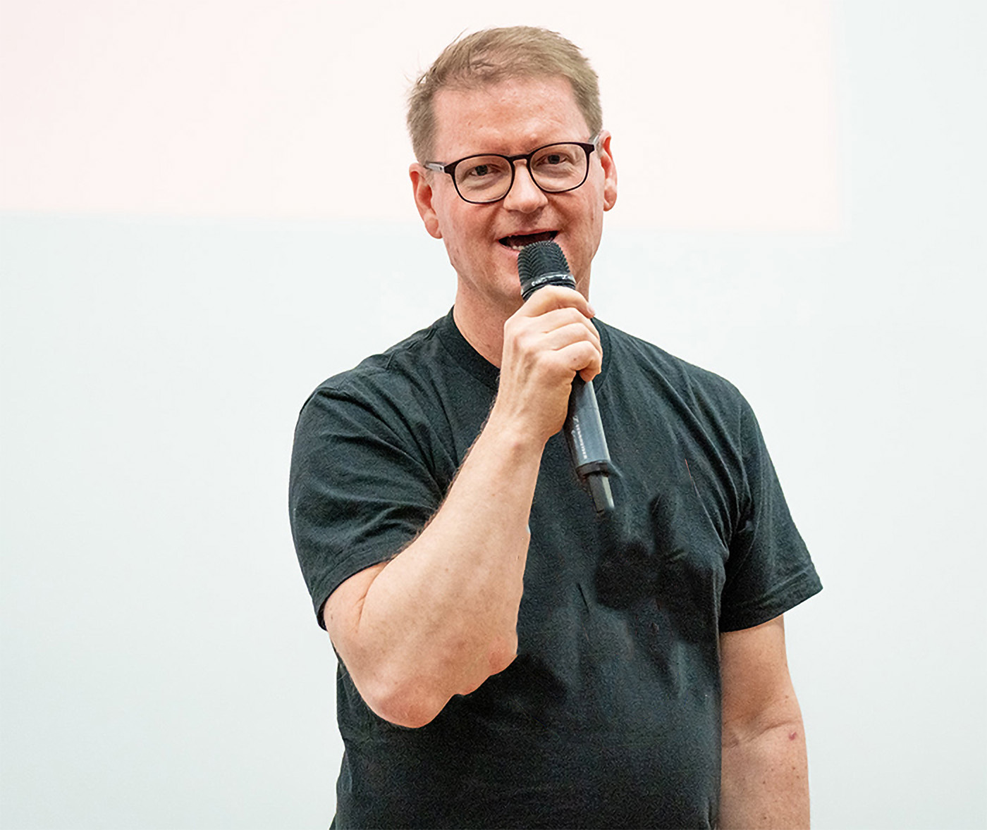 Ein Redner hält ein Mikrofon in der Hand und spricht, während er lächelt. Der Hintergrund ist hell und neutral gestaltet.
