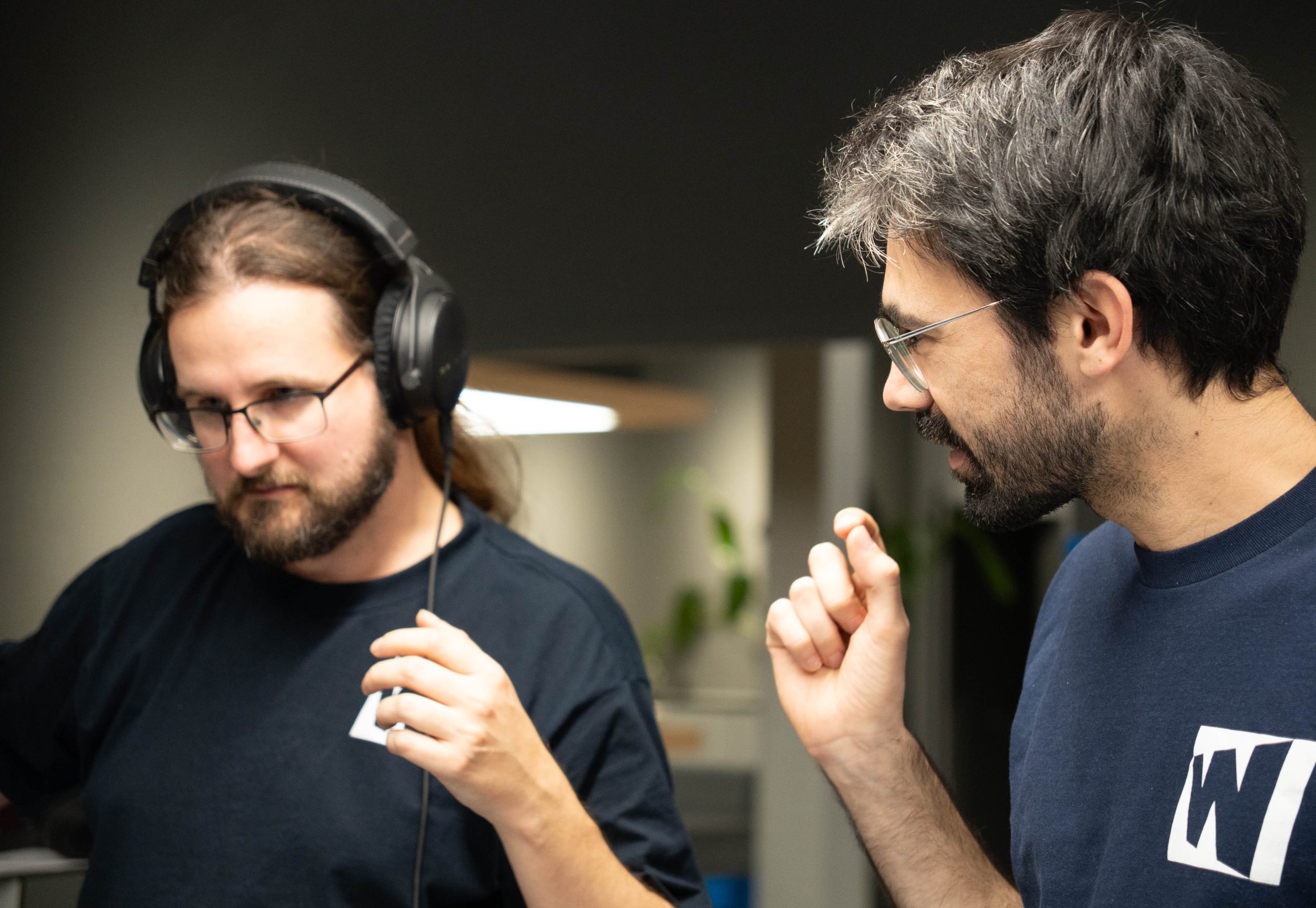 Zwei Männer in T-Shirts stehen in einem Innenraum, einer trägt Kopfhörer und hält seinen Arm, während der andere mit erhobener Hand spricht.